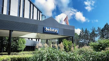 Een modern hotel met een 'Belstay' bord, omgeven door verzorgde groene beplanting en een helderblauwe lucht.