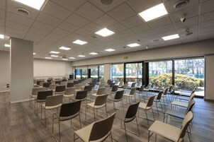 Uma sala de conferências com fileiras de cadeiras e mesas, amplas janelas e uma tela de projeção.