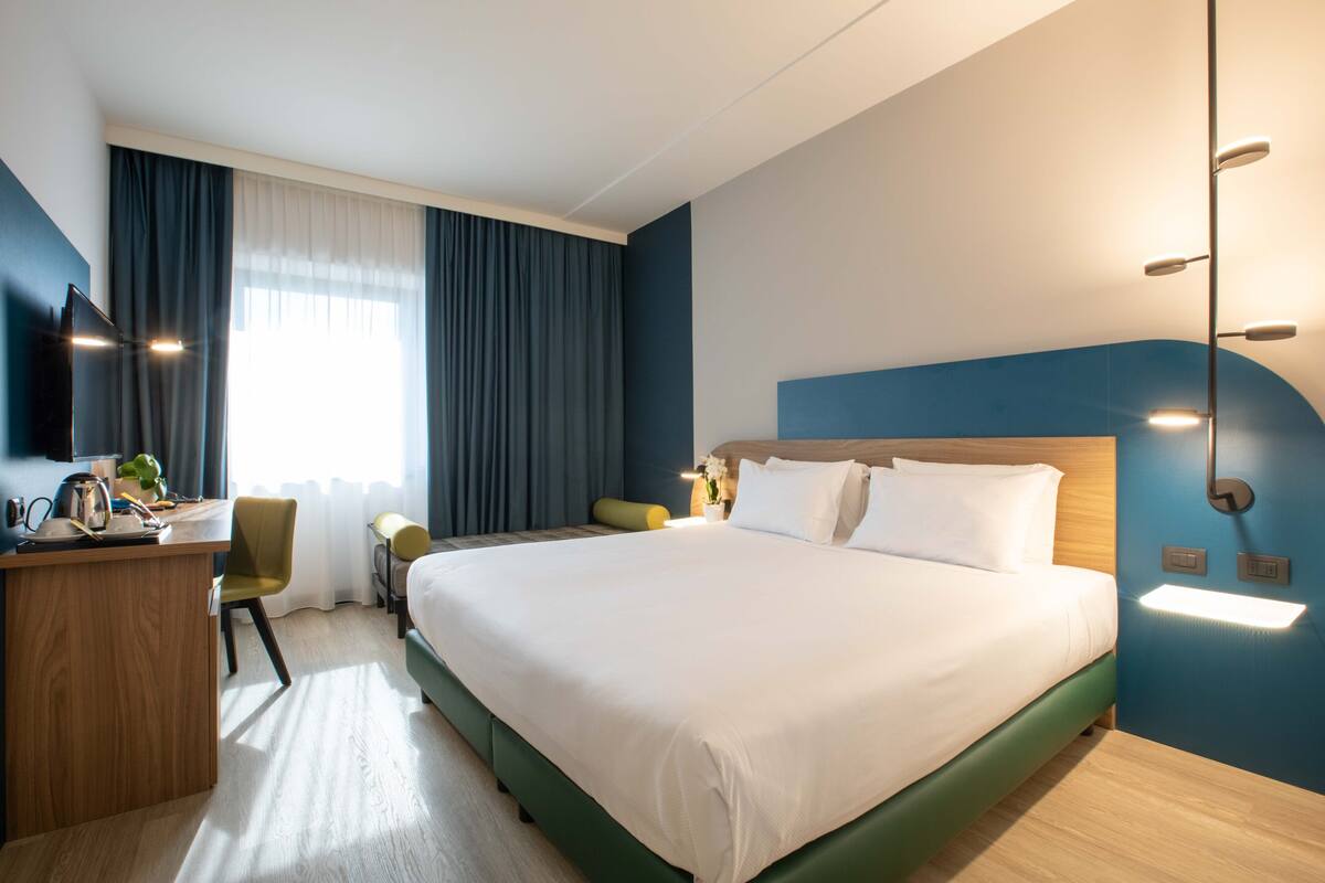 A hotel room with a large bed, a desk, a chair, and a desk lamp.