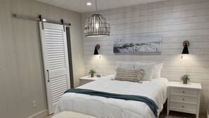 A bedroom with a bed, bedside tables, a bench, a hanging light fixture, and a painting on the wall. - Beulah Getaway (Pensacola)