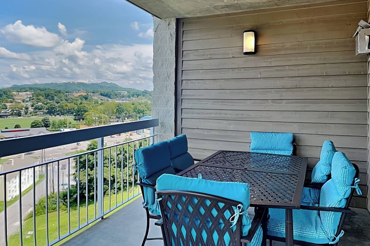 A balcony with a table and chairs, a view of a green landscape and buildings, and a wooden wall.