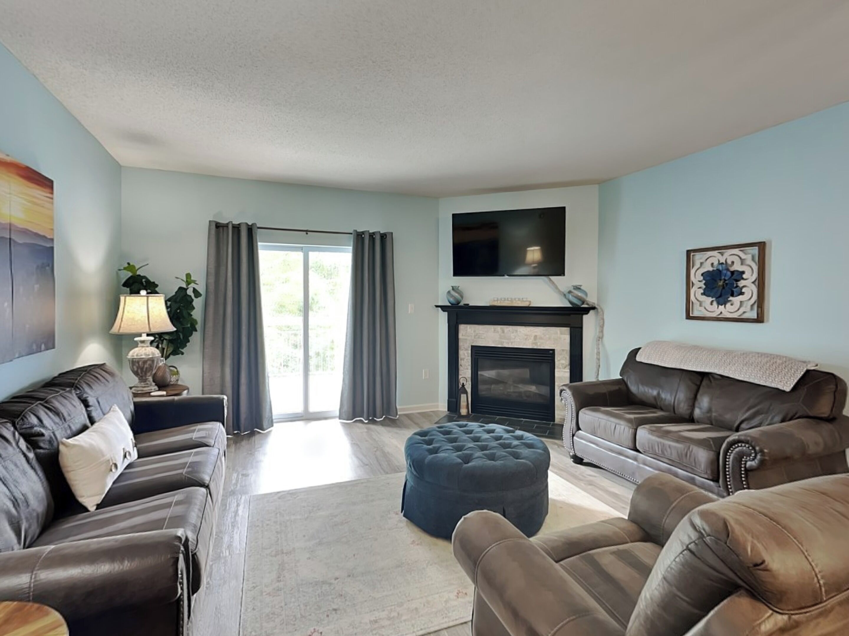 A living room with a fireplace, two leather sofas, a large ottoman, a flat-screen TV, and a sliding glass door leading to a balcony.