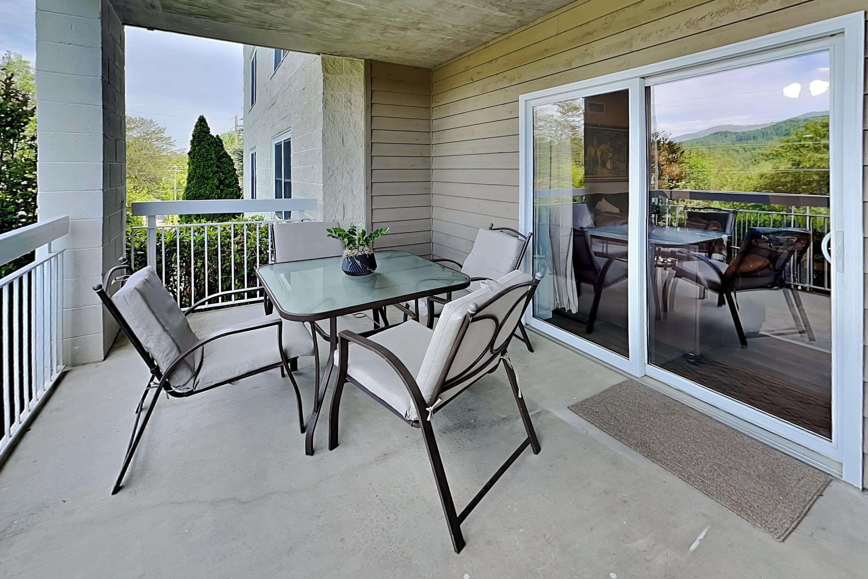 Un patio cubierto con una puerta de cristal que da a una terraza, con una mesa y sillas, una planta en maceta y vistas a una casa y colinas.