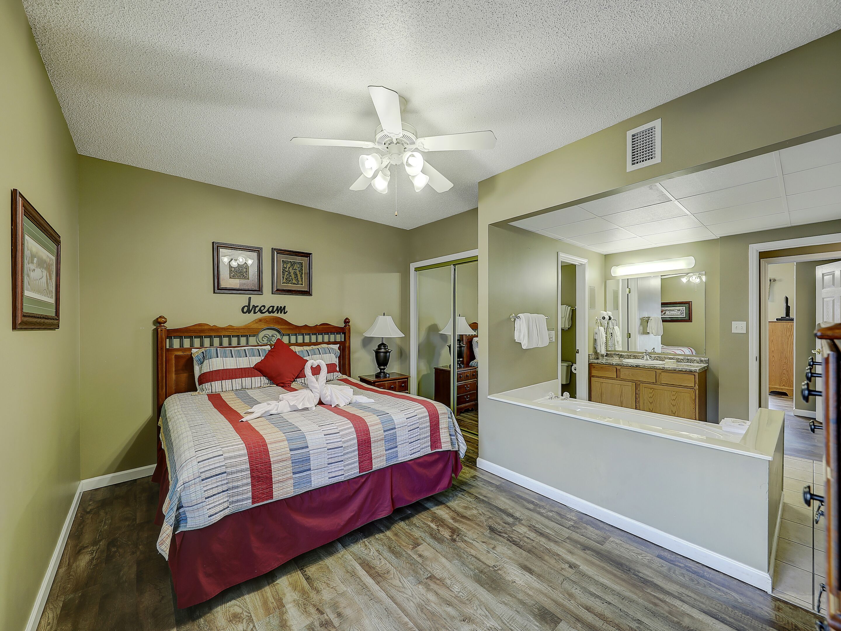 Une chambre à coucher comprenant un lit, un ventilateur de plafond, une table de chevet, une lampe, une commode, un miroir et une salle de bain visible à travers une porte ouverte.