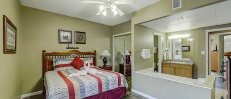 A bedroom with a bed, a ceiling fan, a nightstand, a lamp, a dresser, a mirror, and a bathroom visible through an open door.