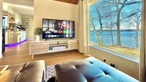 A modern living room with a large flat-screen TV, a comfortable sofa, and a view of a lake through a large window.