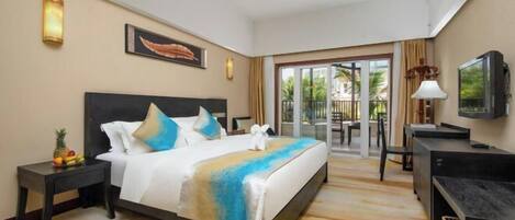 A hotel room with a bed, a desk, a television, and a balcony with a view of palm trees.