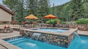 A hot tub with a stone surround, a fire pit, outdoor seating, and two umbrellas. - Ski-in/Out Elegance in Beaver Creek + Fireplace | Chalet Bello (Beaver Creek)