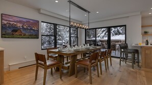 Un'area pranzo con un tavolo di legno apparecchiato per sei persone, ampie finestre con alberi innevati e un dipinto di un paesaggio montano sulla parete.