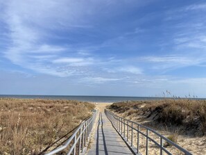 A pathway leading to a beach with a metal railing, sandy dunes, and a clear sky.