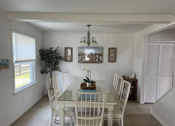 A dining room with a glass table, white chairs, a chandelier, framed artwork, and a potted plant.