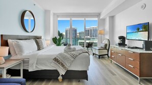A modern hotel room with a large bed, a television, a desk, and a view of the cityscape through large windows. - SF Beautiful Blue & Gold Studio with Ocean View (Miami)