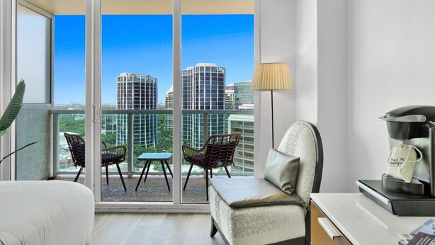 A modern hotel room with a balcony, a sofa, a coffee machine, and a view of city buildings. - SF Beautiful Blue & Gold Studio with Ocean View (Miami)