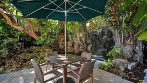 A patio with a table and chairs under a green umbrella, surrounded by lush greenery and a rock waterfall. - SF Ocean View 2BD/2Bth @ Hotel IN Coconut Grove (Miami)