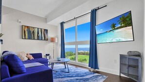 A modern living room with a large window, a blue sofa, a round coffee table, and a flat-screen TV displaying a beach scene. - SF Ocean View 2BD/2Bth @ Hotel IN Coconut Grove (Miami)