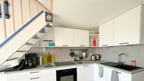 Eine kleine Küche mit weißen Schränken, einem schwarzen Herd und einem Spülbecken. Es gibt eine Kaffeemaschine, einen Wasserkocher und verschiedene Küchenutensilien.