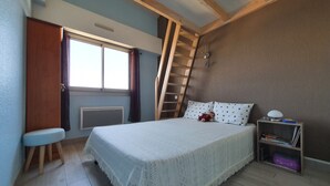 Ein Schlafzimmer mit Bett, Fenster, einer Holzlade, einem Nachttisch und einem Bücherregal.