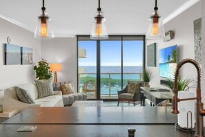 Een ruime woonkamer met een groot raam met uitzicht op zee, een comfortabele bank, een salontafel en een bureau met een lamp.