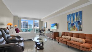 A modern hotel room with a large bed, leather seating, a glass coffee table, a desk with a computer, a TV, and a painting of sailboats. - SF Amazing Studio - Walking Distance to Cocowalk (Miami)