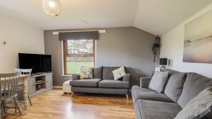 A modern living room with a grey sofa, a dining table with chairs, a flat-screen TV, and a large window with a view of a green landscape. - Pengeulan (Machynlleth)