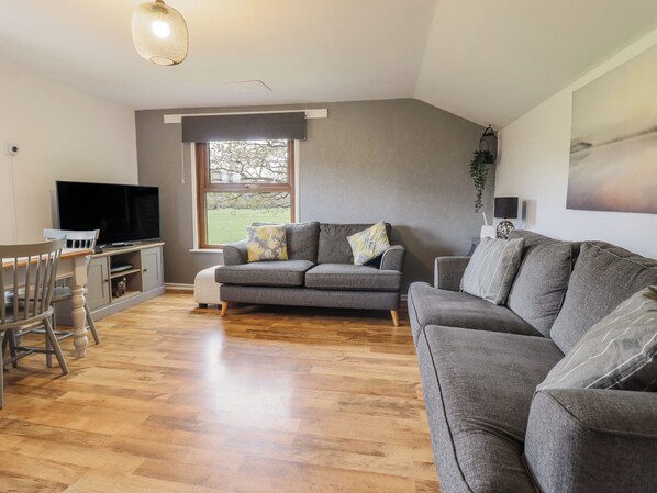 Una sala moderna con un sofá gris, una mesa de comedor con sillas, un televisor de pantalla plana y un amplio ventanal con vistas a un paisaje verde.