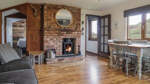 A cozy living room with a brick fireplace, a dining area with wooden furniture, and a view of the outdoors through a large window. - Pengeulan (Machynlleth)