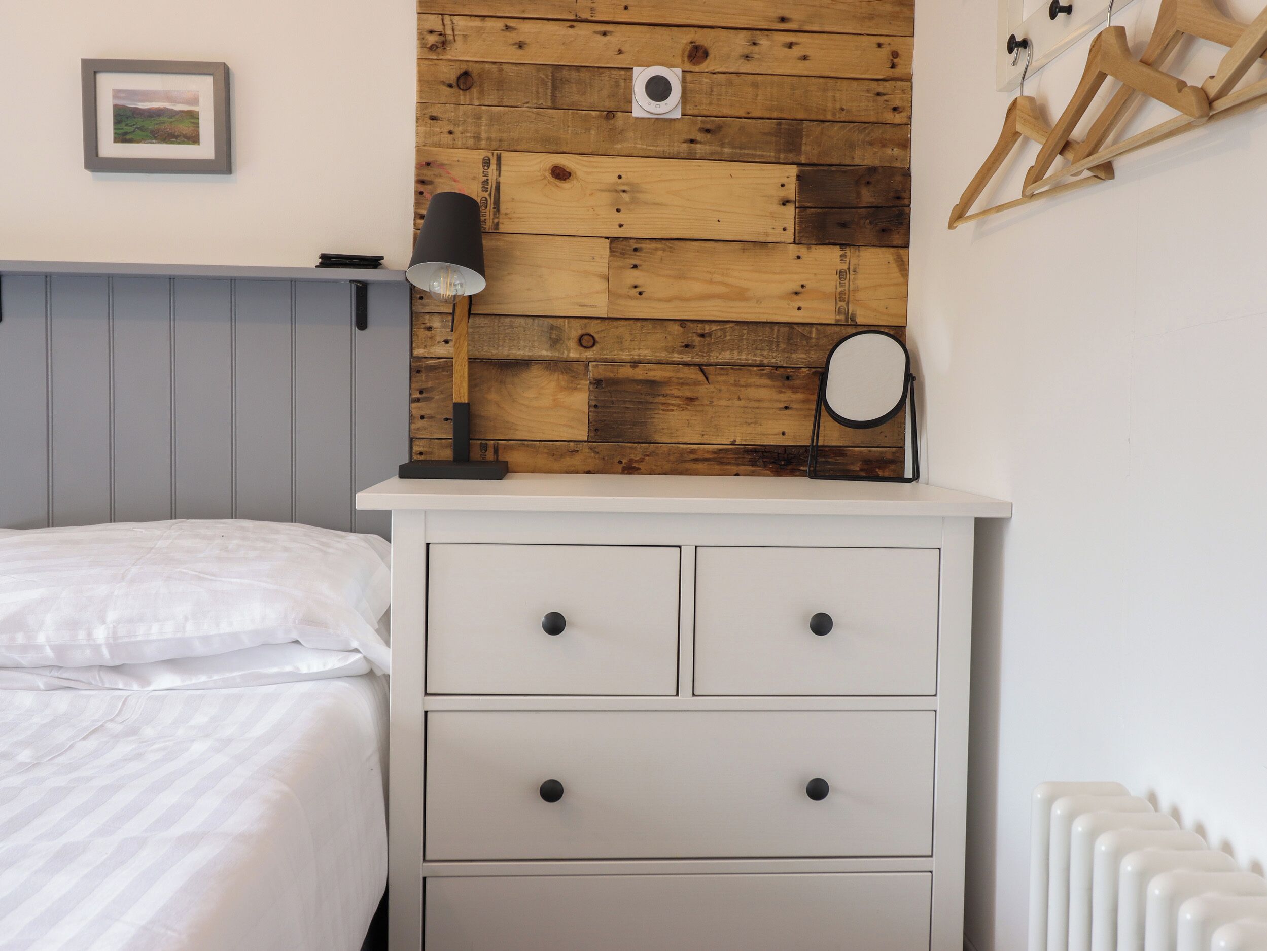 Een slaapkamer met een houten hoofdeinde, een witte ladekast, een bedlampje en een lijst met een afbeelding aan de muur.