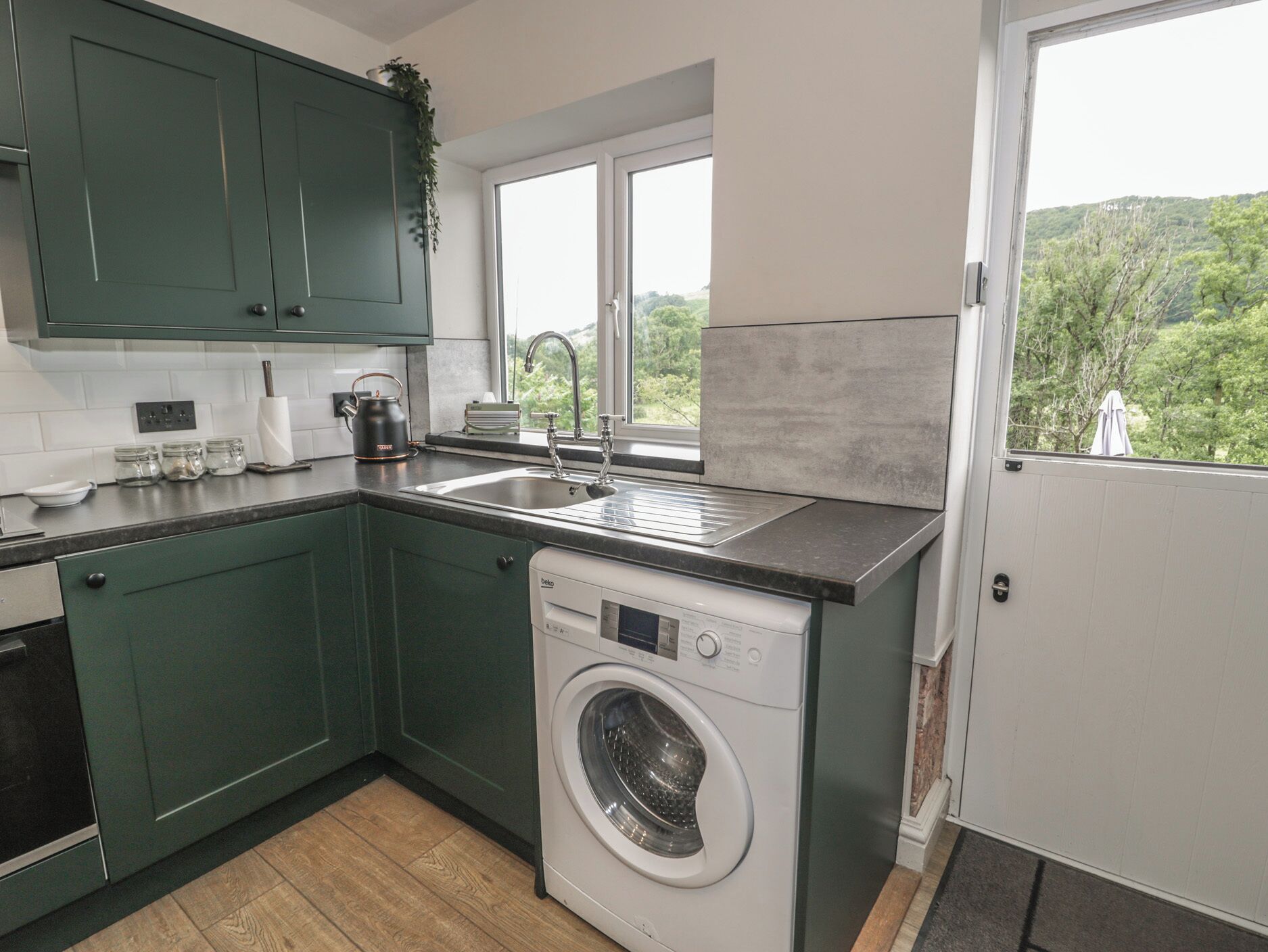 Cocina compacta con lavadora, lavaplatos y ventana con vistas a un paisaje verde.