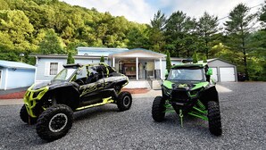 Zwei Geländewagen vor einem Haus mit Kiesweg und einem bewaldeten Hügel im Hintergrund.