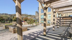 An outdoor patio area with wooden pergolas, stone planters, and a view of a stone building in the background. - Over The Arch (Keighley)