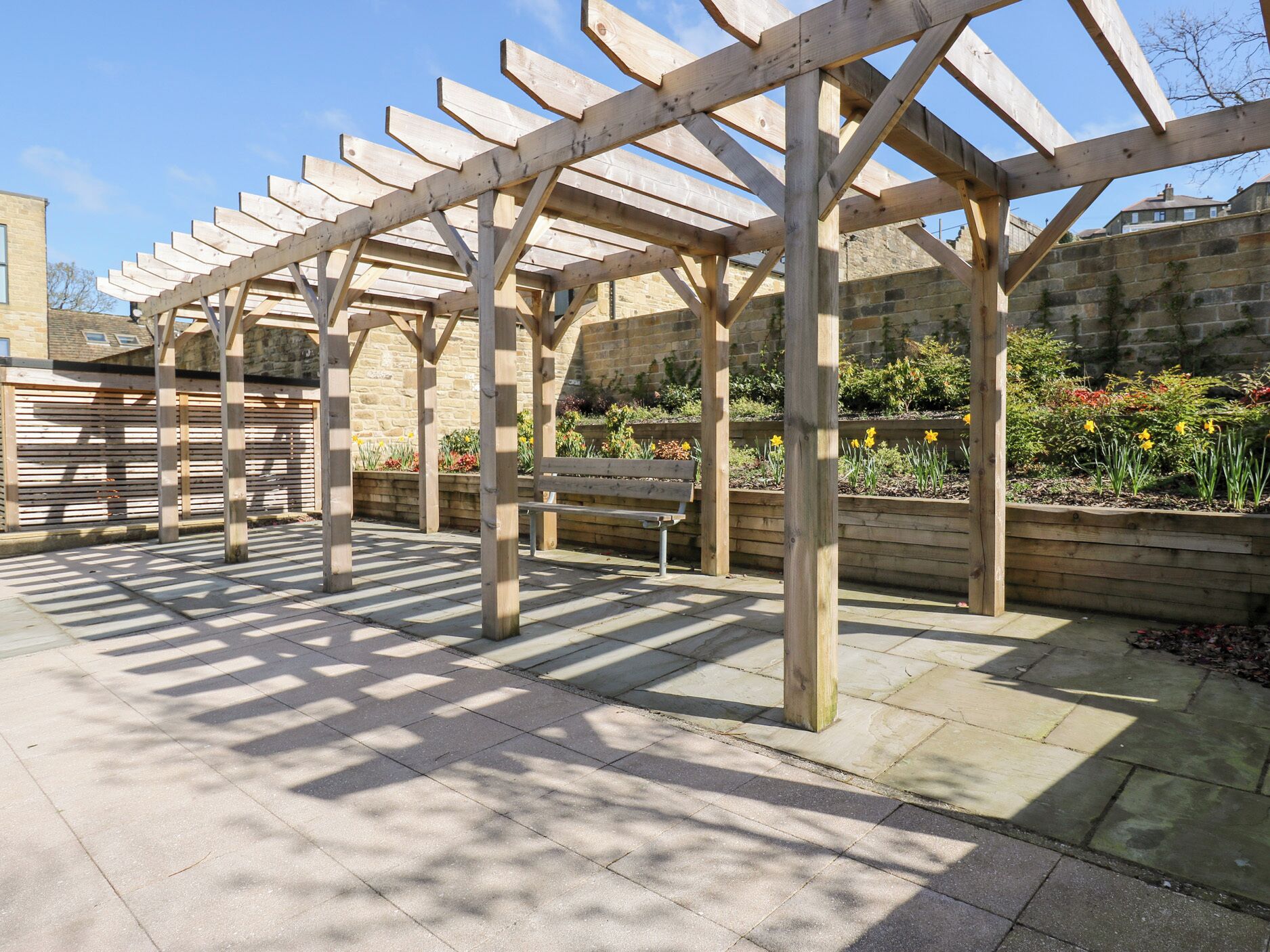 A pergola structure with a bench underneath, set against a stone wall with flowering plants.