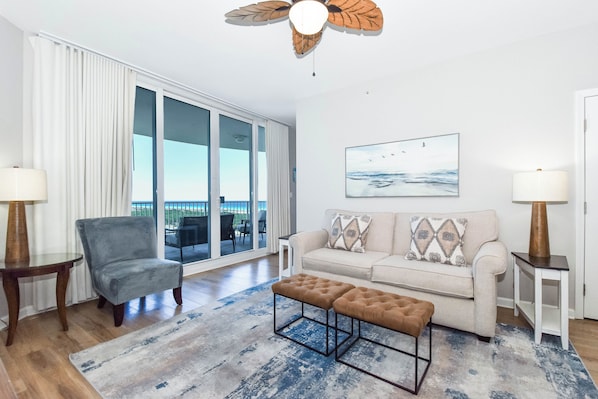 A modern living room with a sofa, ottoman, and chair, a large window with curtains, a ceiling fan, and a painting on the wall. - 11th Floor FULL 2 Bedroom at The Palms of Destin (Destin)