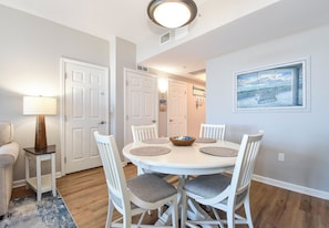 A dining area with a round table, white chairs, a bowl of fruit, and a painting on the wall. - 11th Floor FULL 2 Bedroom at The Palms of Destin (Destin)
