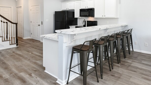 A kitchen with white cabinets, a black refrigerator, a microwave, and a marble countertop. There are five stools with wooden seats and metal frames.