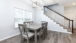 A dining area with a wooden table and chairs, a staircase with white railings, and a window with blinds.