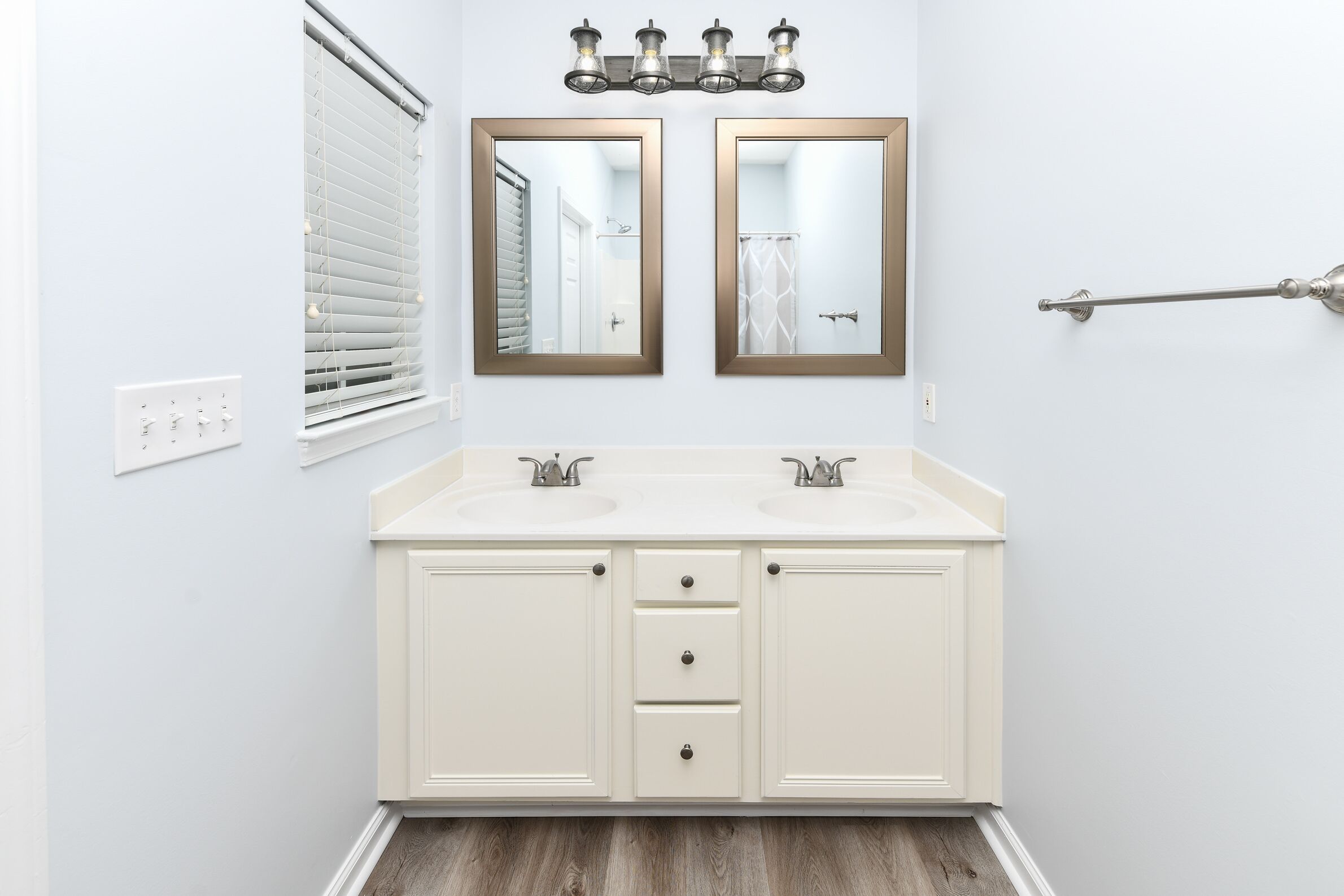 A bathroom with a double vanity, two mirrors, a towel rack, and a window with blinds.
