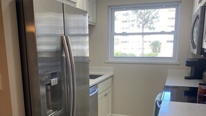 A modern kitchen with stainless steel appliances, white cabinetry, and a window providing natural light. - Beach Front Condo (Bonita Springs)