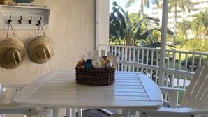 A white table and chairs on a balcony with a view of palm trees and a building. There are two woven hats hanging on hooks and a basket with items on the table. - Beach Front Condo (Bonita Springs)
