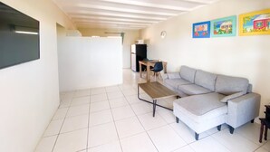 Uma sala de estar com um sofá cinza, uma mesa de centro de madeira e uma TV fixada na parede. Há quadros coloridos na parede e um chão revestido com azulejos.