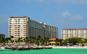 Um hotel grande com vários andares e varandas, uma praia com palmeiras e um cais com barcos.