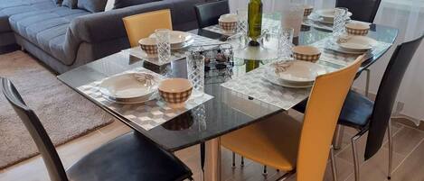A dining area with a glass table set for six, surrounded by modern chairs, a sofa, and a large window.