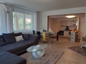 A modern living room with a dark grey sofa, a glass coffee table, and a dining area with a table and chairs. A kitchen is visible in the background with a microwave and a fruit bowl. - Holiday apartment with pool (Malinska)