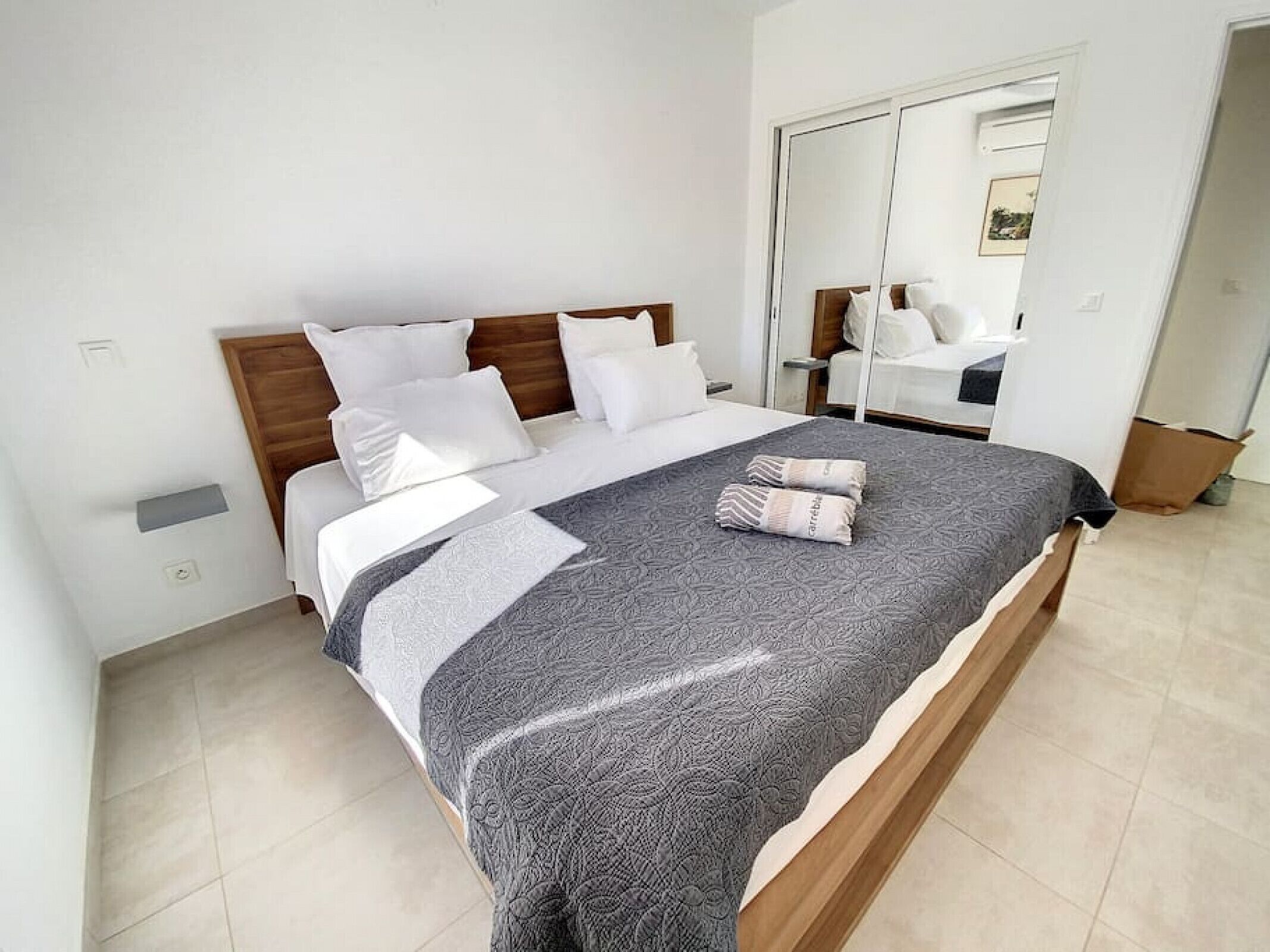 A neatly made bed with a patterned bedspread and pillows, a wooden headboard, and a white wall with a small shelf.
