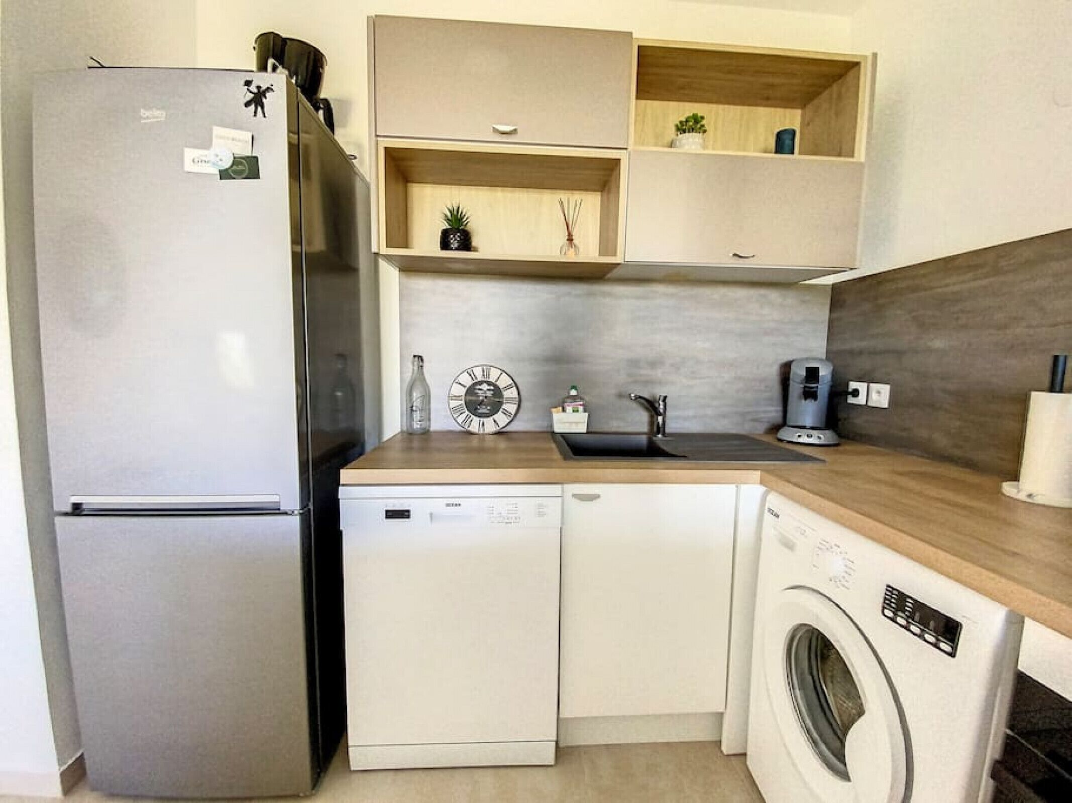 Cocina compacta con refrigerador, lavavajillas y lavadora. Hay estantes de madera sobre el lavabo y un reloj en la pared.