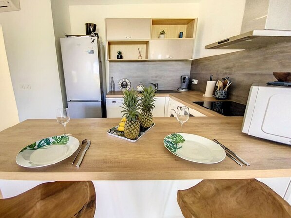 A modern kitchen with wooden countertops, a white refrigerator, and a microwave. There are two dining tables set with plates and cutlery, and a tray of pineapples on one table.