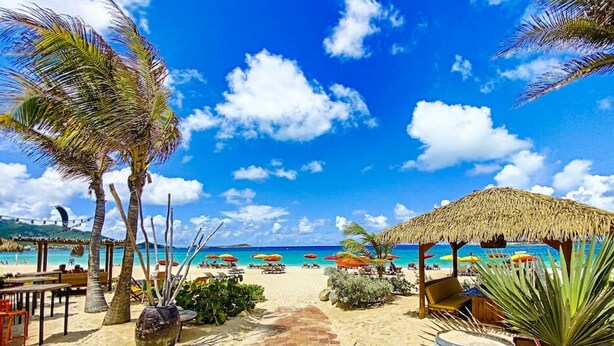 A beachside walkway leading to a thatched-roof structure with outdoor seating and umbrellas.
