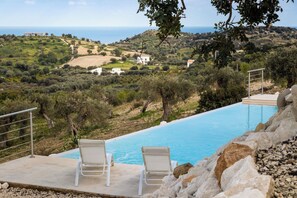 Una piscina con due lettini bianchi, circondata da rocce e ulivi, con vista sul paesaggio costiero.