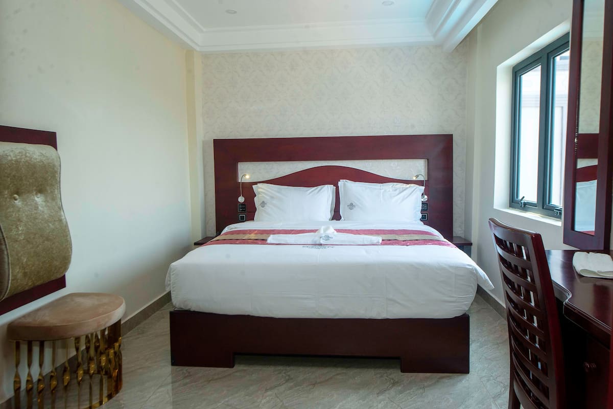 Una cama bien hecha con ropa blanca y un detalle rojo, cabecera de madera y un pequeño taburete al lado.