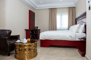 Una habitación de hotel con una cama, un sillón de cuero, una mesita redonda con una tetera y jugo de naranja, y un escritorio con una silla.