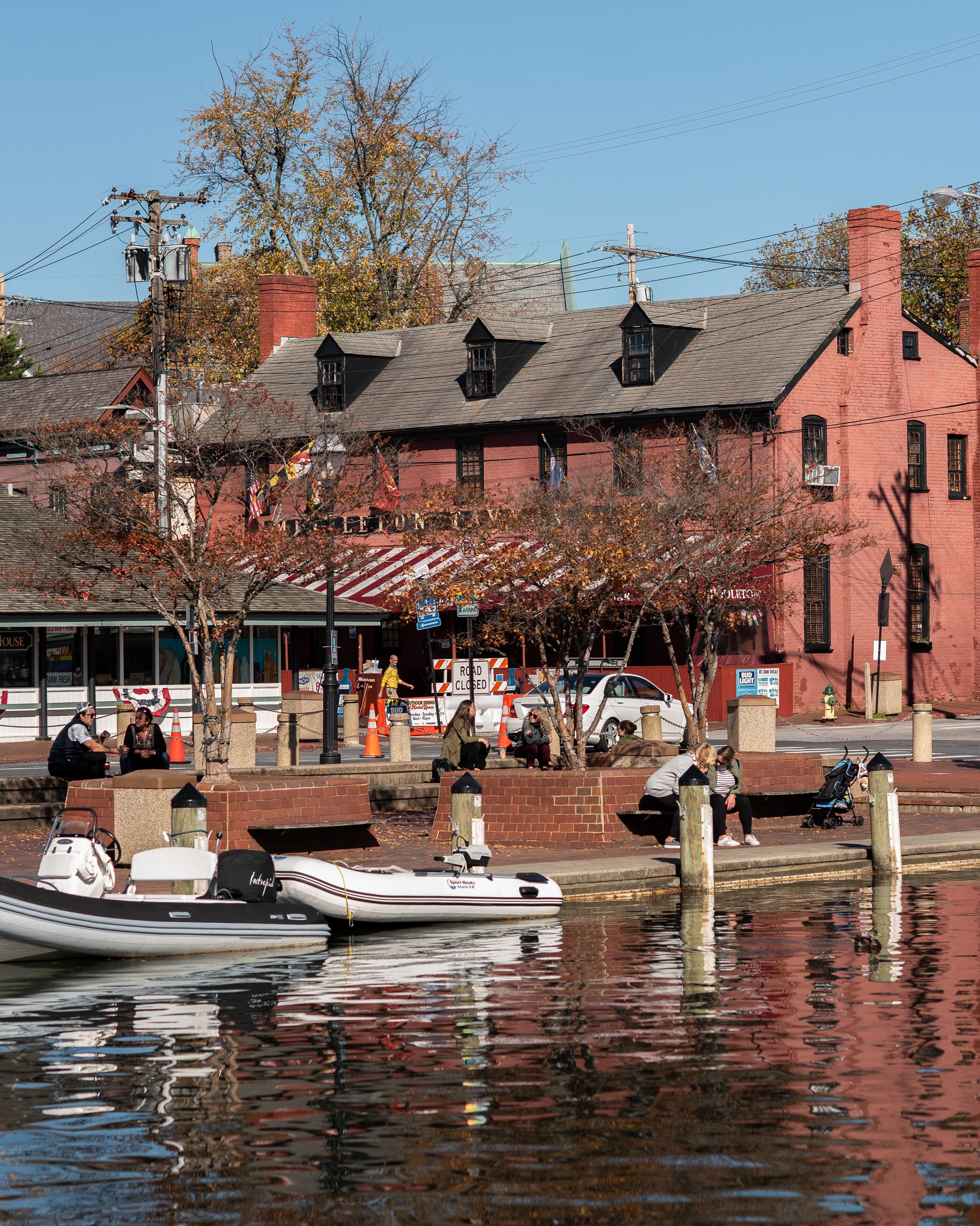 A waterfront scene with a red brick building, a dock with a boat, and people near the water.